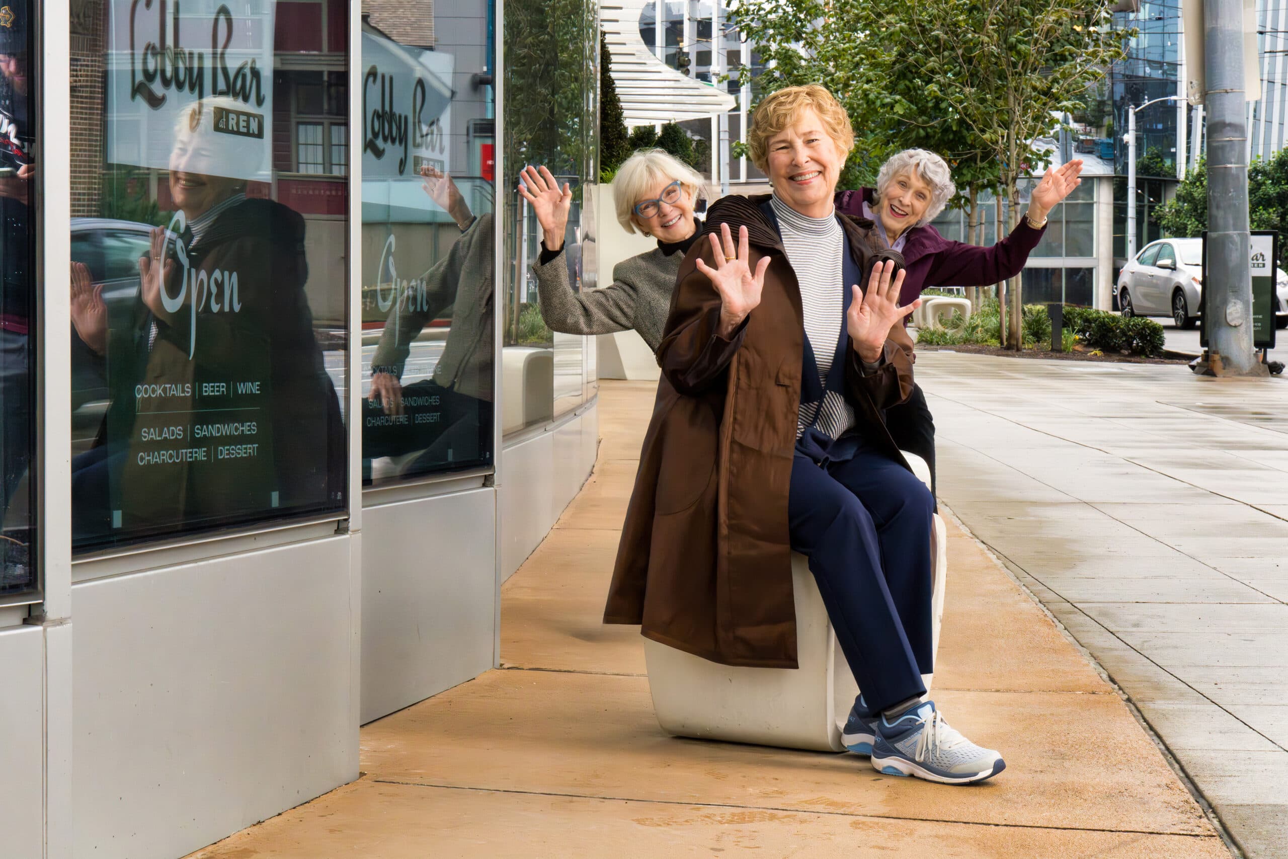 Three smiling women doing a fun pose for a photo outdoors. Their hands are outstretched.